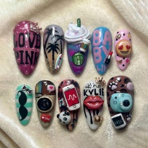 Könnte beinhalten: Ein Set von zehn langen, mandelförmigen Acrylnägeln mit verschiedenen Designs. Die Designs umfassen Text, Emojis und Miniatur-Objekte wie eine Kaffeetasse, Lippenstift und eine Kamera. Farben sind Pink, Blau, Weiß und Braun.