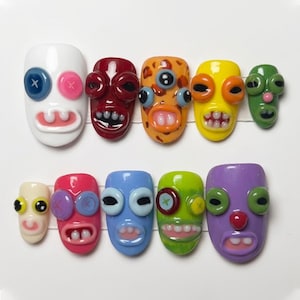 May include: A collection of ten colorful, whimsical nail art designs. Each nail features a unique face design with button eyes, open mouths, and various colors including white, red, orange, yellow, green, pink, blue, and purple. The designs are arranged on a white surface.