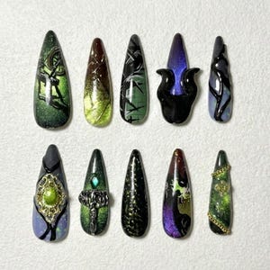 May include: A set of ten long, almond-shaped artificial nails. Each nail features unique designs, including green, black, and purple gradients, intricate patterns, and decorative elements like gold chains and gems. The overall theme is dark and fantasy-inspired.