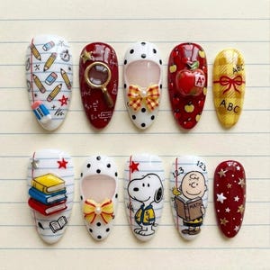 May include: A set of ten press-on nails featuring school-themed designs including pencils, erasers, a magnifying glass, math formulas, apples, a yellow plaid pattern with ABC text, stacked books, Snoopy, Charlie Brown, and star accents.