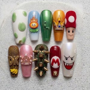 May include: A set of ten manicured fingernails with various designs. The designs include a Yoshi egg, a fire flower, the letter "L", a gold crown, a mushroom, a Donkey Kong design, a pink nail with a crown, a spiky green shell, a Mario symbol, and a Boo ghost.