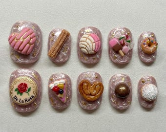 May include: A set of ten artificial nails with a shimmering pink base, adorned with miniature food-themed decorations. Designs include a heart, churro, concha, popsicle, donut, slice of cake, palmiers, and a De La Rosa rose logo.