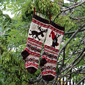 Peut inclure: Deux bas de Noël tricotés avec des motifs marron, rouges et crème, comprenant des rennes et des motifs géométriques. Les bas sont suspendus à une branche d'arbre, mettant en valeur un thème de vacances festif.
