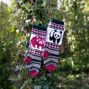 May include: Two knitted Christmas stockings with a red and white panda bear on one and a red and white bear on the other. The stockings are hanging on a tree branch with green leaves.