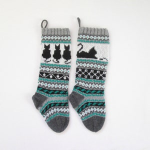 May include: Two gray knitted Christmas stockings with black cat designs. One stocking features three black cat silhouettes, and the other has a single cat. Both stockings have teal, black, and white geometric patterns.
