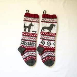 May include: Two knitted Christmas stockings with a red, white, and gray pattern. Each stocking features a dog silhouette and snowflake designs. The top and heel are dark gray, and the loop is red.