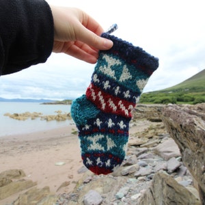 May include: A hand holds a knitted Christmas stocking with a blue, white, red, and black geometric pattern.