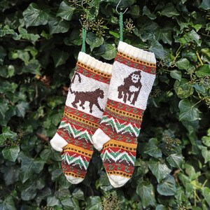 Puede incluir: Dos calcetines de Navidad tejidos, con diseños de animales marrones sobre fondo blanco y patrones geométricos en rojo, verde y dorado. Los calcetines cuelgan sobre un fondo de follaje verde, creando un ambiente festivo navideño.