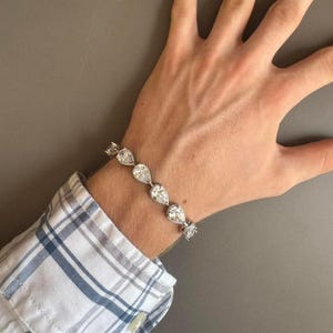 Puede incluir: Una pulsera de plata con una serie de piedras preciosas claras en forma de pera. La pulsera se lleva en la muñeca, con una camisa a cuadros visible en el fondo. Las piedras están engastadas en plata, creando un accesorio brillante y elegante.