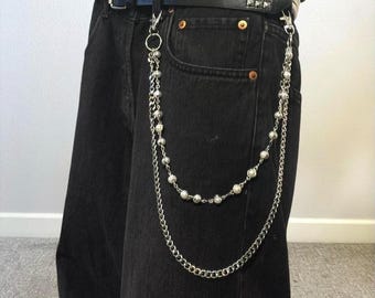 Può includere: Accessorio per cintura a catena argentata con due fili, uno con piccole perle e l'altro con una catena a maglie. La catena è attaccata a una cintura nera con borchie argentate e un paio di jeans neri.
