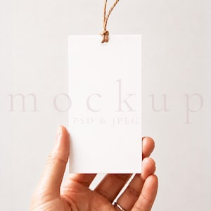 Op de afbeelding: Een blanco witte rechthoekige tag met een bruin touwtje aan de bovenkant. De tag wordt door een hand tegen een witte achtergrond gehouden. Het woord "mockup" is vaag zichtbaar op de tag.