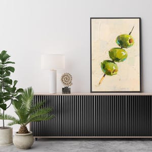 May include: An abstract painting of three green olives on a skewer against a cream-colored background. The artwork is framed in a black rectangular frame and is displayed on a black cabinet with a white lamp and decorative items.