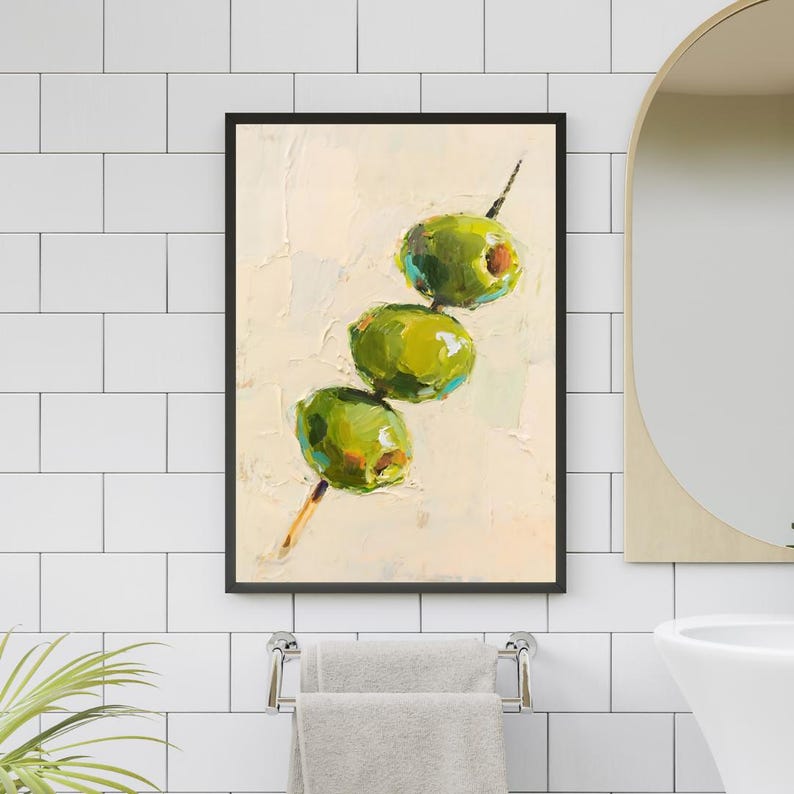 May include: A framed painting depicts three green olives on a skewer against a cream-colored background. The artwork uses thick brushstrokes to create a textured effect, with the olives showcasing highlights of yellow and orange.