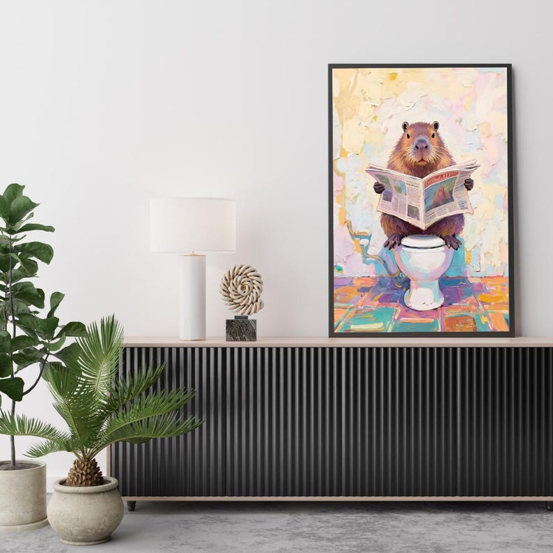May include: A framed art print depicts a capybara sitting on a toilet, engrossed in a newspaper. The artwork features a vibrant, colorful palette with a textured, painterly style. The capybara is brown, and the toilet is white. The background has a mix of yellow, pink, and blue.