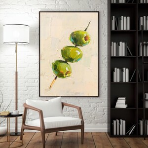 May include: An oil painting depicts three green olives on a wooden skewer against a textured off-white background. The olives have a vibrant green hue with hints of yellow and blue, and the skewer is a light brown color. The artwork is framed in a dark brown frame.