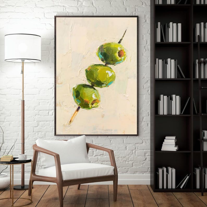 May include: An oil painting depicts three green olives on a wooden skewer against a textured off-white background. The olives have a vibrant green hue with hints of yellow and blue, and the skewer is a light brown color. The artwork is framed in a dark brown frame.