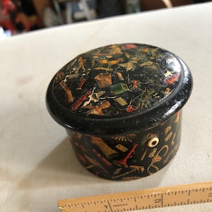 May include: A small, round, black box with a hinged lid. The box is decorated with a colourful pattern of various objects, including dice, playing cards, and musical instruments. The box is approximately 2.5 cm in diameter.
