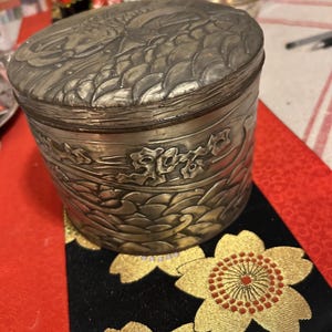 May include: A silver metal container with an ornate design. The container has a lid and is decorated with a floral pattern. The container is sitting on a black and gold fabric with a floral pattern.