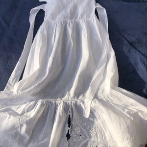May include: A white cotton baby dress with a scalloped hem and embroidered details. The dress has a split back and ties at the shoulders.