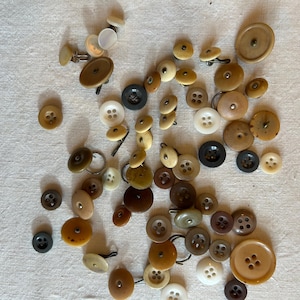 Peut inclure: Une collection de boutons ronds assortis dans diverses nuances de brun, beige, crème, blanc, gris et noir. Les boutons varient en taille et présentent différentes configurations de trous, y compris des modèles à deux et quatre trous, certains fixés sur des tiges métalliques.