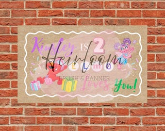 May include: A rectangular banner with a brown background and a white scalloped border. The banner features the text "Kinley is 2 Heirloom Loves You!" with cartoon characters and gift illustrations. The banner is set against a red brick wall.