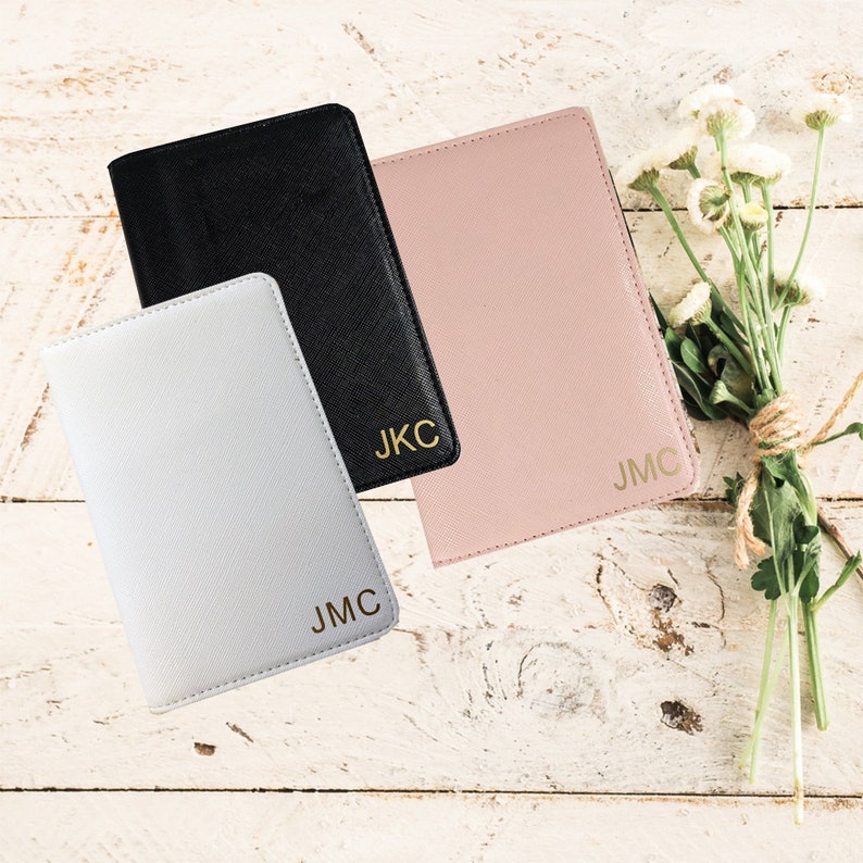 May include: Three passport holders in black, white, and pink with gold lettering. The black passport holder has the initials "J.K.C." on the front. The white passport holder has the initials "J.M.C." on the front. The pink passport holder has the initials "J.M.C." on the front.