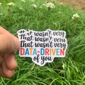 May include: A white sticker with the text "That wasn't very DATA-DRIVEN of you" in a colorful font. The sticker is held in front of a background of green grass and white flowers.