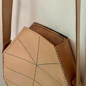 May include: A geometric-shaped, light tan leather bag with a long strap. The bag features green stitching that creates a geometric pattern across the front. The bag's interior is visible, revealing a darker brown lining.