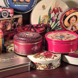 May include: A collection of vintage metal tins in various shapes and sizes. The tins feature colorful designs, including floral patterns, a race car, and a Coca-Cola jukebox. Some tins are round, others rectangular.