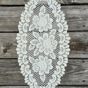 May include: An oval, off-white lace doily with a floral pattern. The doily features three large floral designs and a delicate, openwork mesh. The intricate lace is set against a rustic wooden background.