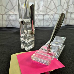 May include: Two clear acrylic clips with metallic silver accents. One clip stands vertically, while the other lies horizontally, holding a bright pink square note and a pale yellow square note. The clips feature a spring-loaded mechanism.