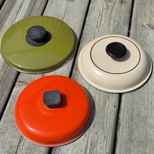 May include: Three round pot lids with black knobs. One lid is olive green, one is bright orange, and one is cream-colored with a thin brown circular accent line. Each lid features a smooth, glossy finish and a simple, functional design.