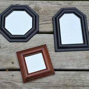 May include: Three decorative mirrors with different frame shapes. One is an octagon with a black frame, another is a rectangle with a black frame, and the third is a square with a brown frame. The mirrors are on a wooden surface.