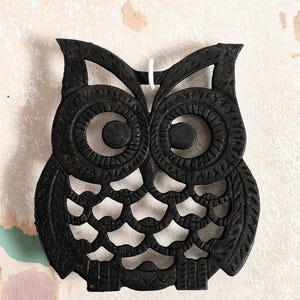 May include: Black cast iron owl-shaped trivet with a detailed, openwork design. The trivet features large, circular eyes and a textured surface. A white string is attached at the top for hanging. This decorative item is suitable for kitchen use.