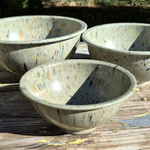 May include: Three round bowls with a light beige base color featuring a speckled pattern of small blue, orange, yellow, and black flecks. The bowls have a smooth, curved interior and a wide rim, arranged in a group on a textured surface.