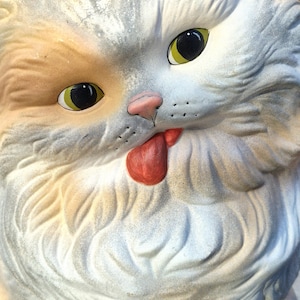 May include: A close-up of a ceramic cat statue with a surprised expression. The cat has white, tan, and gray fur, with large yellow eyes and a pink nose. Its red tongue is sticking out. The statue has a vintage, weathered appearance.