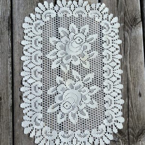 Puede incluir: Un tapete de encaje rectangular de color blanco roto que presenta un patrón floral repetitivo en el centro y un borde festoneado decorativo. El intrincado trabajo de aguja muestra un fondo de malla con motivos florales detallados.