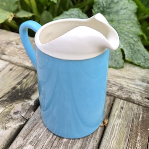 May include: A ceramic pitcher featuring a smooth, light blue cylindrical body and a contrasting white rim with a prominent pouring spout. The pitcher includes a matching light blue curved handle attached to the side, with a clean, glossy finish.