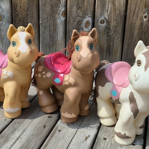 May include: Three plastic toy horses with molded manes and tails. The left horse is tan with a white blaze and pink saddle. The center horse is brown with white spots and a pink saddle. The right horse is white with brown patches and a pink saddle.