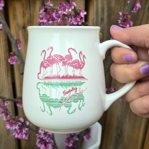 May include: White ceramic mug featuring a pink flamingo design with the text "Siesta Key Florida" in green and pink. The mug has a curved handle and is held against a wooden background.