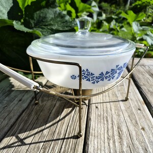 May include: A white glass casserole dish with a blue floral pattern and a clear glass lid. The dish is held in a gold-toned metal warming stand featuring white plastic handle grips and a central base component for a heating element.