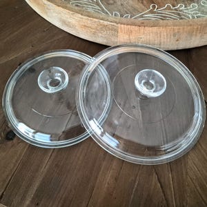 May include: Two circular, transparent glass lids for cookware, each featuring a clear, centered knob handle. The lids have a smooth, flat surface with a slightly raised outer rim, designed to fit securely onto pots or pans for cooking purposes.