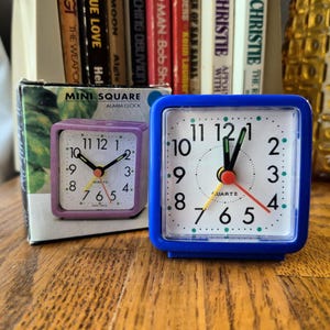 May include: A blue square analog alarm clock with a white face, black numerals, and green dot markers. The clock features black hour and minute hands, a red second hand, and a yellow alarm hand. The packaging box displays a purple version of the clock.