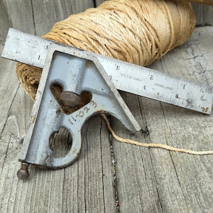May include: A vintage combination square and ruler with visible wear, resting on a weathered wooden surface. A roll of tan twine is in the background. The ruler is marked with inches and the square has the number "11-023" on it.
