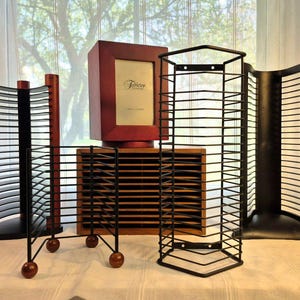 May include: A collection of CD and DVD storage units in various styles. Includes a red wooden photo frame, a black metal rack, and wooden and black storage racks. The items are arranged on a white surface in front of a window.