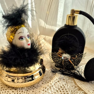 May include: Two vintage perfume bottles. One is a black bottle with a gold-colored top and a black bulb atomizer. The other is a gold-colored container with a doll head on top, adorned with sequins and black feathers.