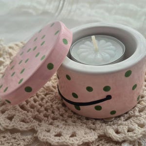 May include: A pink ceramic candle holder with a lid, decorated with green polka dots. The lid is slightly ajar, revealing a white tea light candle inside. The holder sits on a crocheted doily.