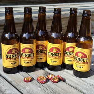 May include: Six amber glass bottles of Sunset Sarsaparilla soda from Jones Soda Co. featuring yellow labels with red text and a barrel graphic. Four red metal bottle caps with the Sunset Sarsaparilla logo are placed in front of the bottles.