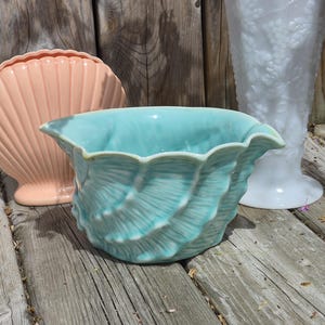 May include: A turquoise ceramic seashell-shaped bowl with a scalloped rim. A peach-colored seashell-shaped vase and a white vase with a grape design are in the background. The objects are on a weathered wooden surface.