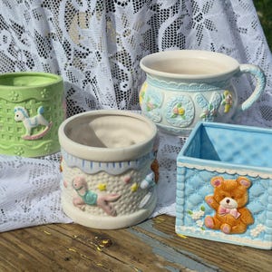 May include: A collection of four ceramic containers featuring pastel colors and raised relief designs. One is a light green cylinder with rocking horses, a cream carousel pot, a light blue mug with floral accents, and a square blue bear planter.